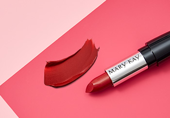 Červená rtěnka Mary Kay vyfotografovaná s uzávěrem položeným nahoře na růžovém pozadí vedle ukázky barvy na rty.