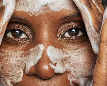 Detailní pohled na zralou ženu myjící si rukama obličej, po celém obličeji – na tvářích, nose a čele – má bílou mýdlovou pěnu.