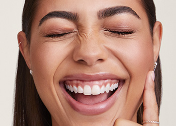 Mladá žena s výrazným obočím a dlouhými rovnými hnědými vlasy, směje se, oči má zavřené a nos pokrčený. 